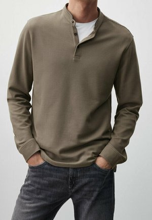 Mand iført olivengrøn langærmet henley-skjorte med to knapper åbne, hænderne i lommerne og mørkegrå jeans.