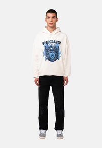 Felpa crema con stampa grafica blu e jeans neri. La felpa ha una vestibilità morbida con cappuccio regolabile. I jeans presentano dettagli strappati.