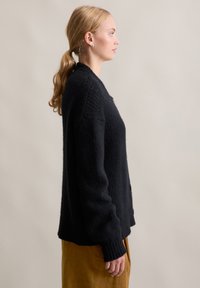 Cardigan nero lavorato a maglia con finitura testurizzata, caratterizzato da maniche lunghe, vestibilità ampia e polsini a coste. Indossato sopra una gonna color senape.