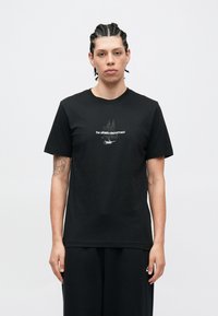 Μαύρο βαμβακερό μπλουζάκι με κοντά μανίκια, που έχει λευκά γράμματα στο κέντρο, φορεμένο με άνετο μαύρο παντελόνι. Κλασικός στρογγυλός γιακάς.