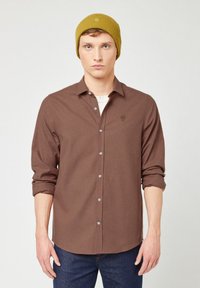 Camicia a scacchi marrone con maniche lunghe, colletto button-down e ricamo sul petto. Abbinata a un berretto di lana giallo. Jeans in denim blu.