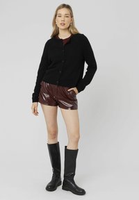Cardigan noir en tricot, shorts bordeaux brillants et bottes noires à hauteur de genou. Design simple avec des boutons sur le cardigan et une texture lisse.