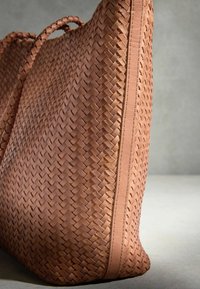Sac à main en cuir tressé marron avec des poignées tressées, posé verticalement sur un fond gris neutre.