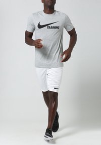 Camisa de entrenamiento Nike en gris con logo negro, combinada con pantalones cortos blancos que tienen un pequeño logo negro, usados con zapatillas deportivas negras.