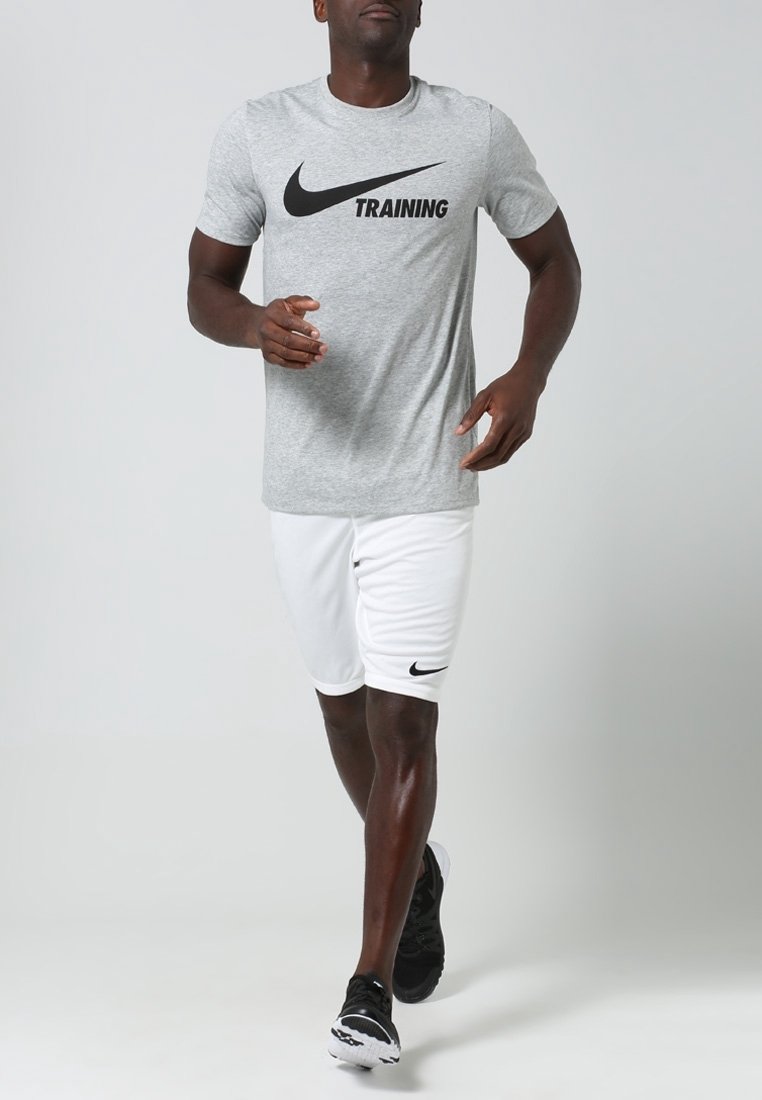 Camisa de entrenamiento Nike en gris con logo negro, combinada con pantalones cortos blancos que tienen un pequeño logo negro, usados con zapatillas deportivas negras.