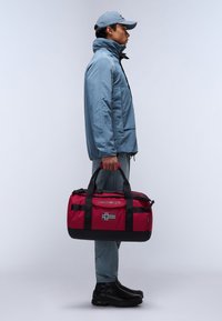 Sac de voyage rouge avec des accents noirs et un détail de drapeau norvégien. Le sac a des poignées doubles et une fermeture éclair, fabriqué à partir de matériaux durables.