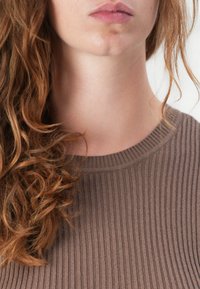 Maglione a coste marrone con scollo a girocollo. Capelli ricci parzialmente visibili, che evidenziano la texture del tessuto e il design aderente.