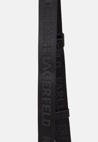 Cinghia in tessuto intrecciato nero con testo "Karl Lagerfeld" in rilievo ripetuto e un piccolo anello in pelle nera su sfondo bianco.