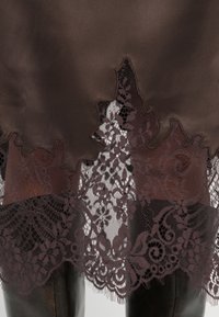 Tissu en satin marron avec des bords festonnés, présentant des détails en dentelle bordeaux complexes. La texture est lisse avec un effet de superposition.