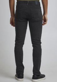 Jeans negros ajustados con una textura de denim suave, con bolsillos traseros y un sutil parche con un logo. Combinados con zapatillas oscuras.