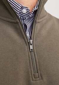 Grijze zip-up trui met een fleece-structuur, voorzien van een zilveren rits. Daaronder is een gekleed overhemd met blauw-witte strepen zichtbaar.