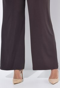 Wijd uitlopende, donkergrijze broek gemaakt van zachte stof met een gladde textuur en een subtiele glans. Draagt nude pumps met hoge hakken.