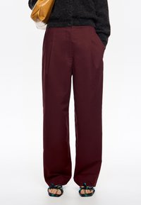 Pantalones de pierna ancha burdeos con una textura suave, que presentan pliegues en la parte delantera y bolsillos laterales, combinados con sandalias coloridas y adornadas.