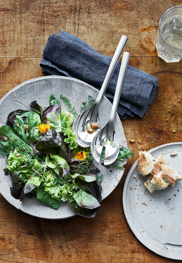 Salade verte mélangée fraîche avec des fleurs comestibles sur une assiette en céramique, des ustensiles à salade en métal, du pain déchiré sur une assiette à côté, et un verre d'eau sur une table en bois.