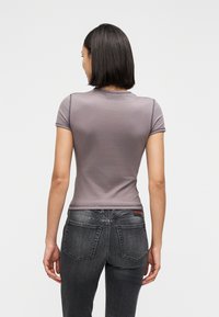 Γυναίκα με σκούρα μαλλιά μήκους μέχρι τους ώμους, φοράει εφαρμοστό γκρι κοντομάνικο πουκάμισο και μαύρα ξεθωριασμένα τζιν, στέκεται πλάτη σε λευκό φόντο.