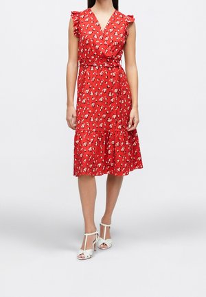 Femme portant une robe portefeuille rouge à fleurs avec des manches volantes et des sandales blanches à talons hauts, debout sur un fond clair uni.