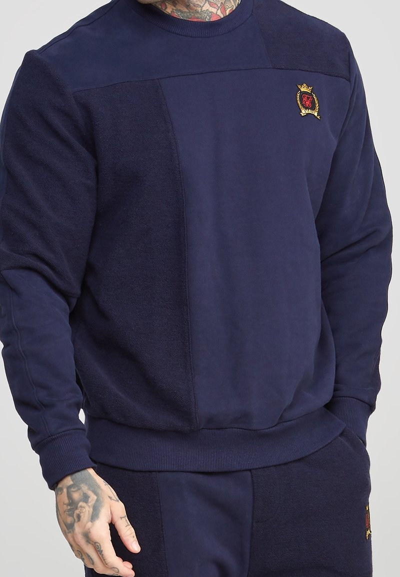 Sweatshirt bleu marine avec panneaux texturés, poignets et encolure côtelés, et logo brodé sur la poitrine. Tissu doux avec un léger éclat.