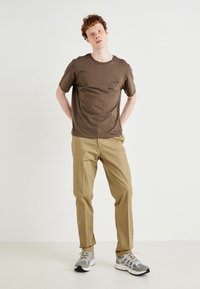 Brown cotton t-shirt with short sleeves and a round neckline. Features a stitched design on the chest. Paired with beige pants and gray sneakers.