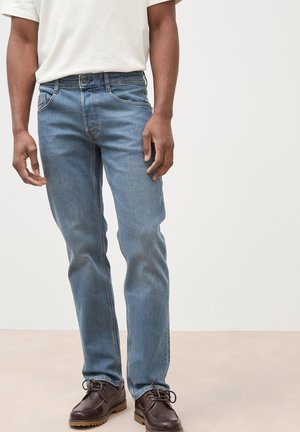 Mann trägt hellblaue Slim-Fit-Jeans, weißes T-Shirt und dunkelbraune Schnürschuhe, steht vor neutralem Hintergrund.
