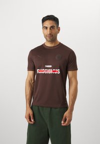 Smeđi kratkodlaki T-shirt s okruglim izrezom, s logotipom na prsima i bijelim tekstom koji glasi "KEIN PLATZ FÜR RASSISMUS", s glatkom teksturom.