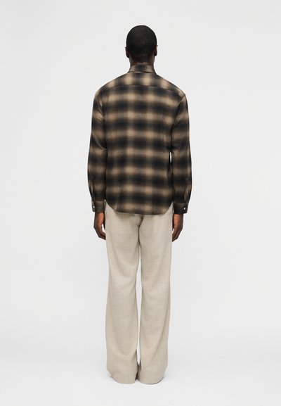 Άνδρας με την πλάτη προς το φακό, φοράει καφέ και μαύρο καρό πουκάμισο με μπεζ παντελόνι φαρδιάς γραμμής, μπροστά σε απλό φωτεινό φόντο.