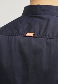 Námořnická modrá košile s hladkým povrchem, s klasickým límečkem a oranžovou visačkou na zadní straně krku. Jednoduchý design, bez vzorů nebo akcentů.