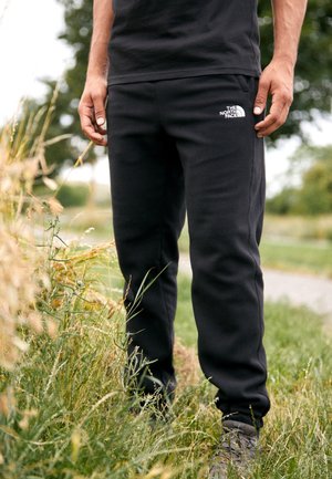 Mand iført sorte The North Face joggingbukser og mørke sko står på græs nær en jordsti med grønne træer i baggrunden.