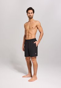 Schwarze Badshorts mit einem elastischen Bund, seitlichen Taschen und einem kleinen Logo am linken Saum. Das Modell steht barfuß vor einem neutralen Hintergrund.