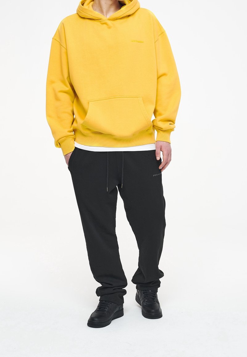 Personne portant un sweat à capuche jaune avec une poche frontale, pantalon noir à cordon, baskets noires, debout avec une main dans la poche.