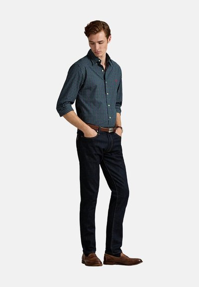 Camisa de cuadros verde y azul marino con botones, mangas largas y cuello, combinada con jeans oscuros y zapatos marrones, con un corte clásico y detalles sutiles.