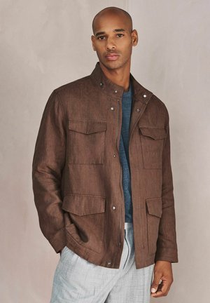 Man wearing a brown jacket with front pockets, a blue shirt, and light gray pants, standing against a plain beige background.