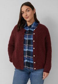 Bordeauxfarbener Strickcardigan mit einer weichen Textur, runder Halslinie und weißen Knöpfen. Über einem blau-schwarz karierten Hemd getragen.
