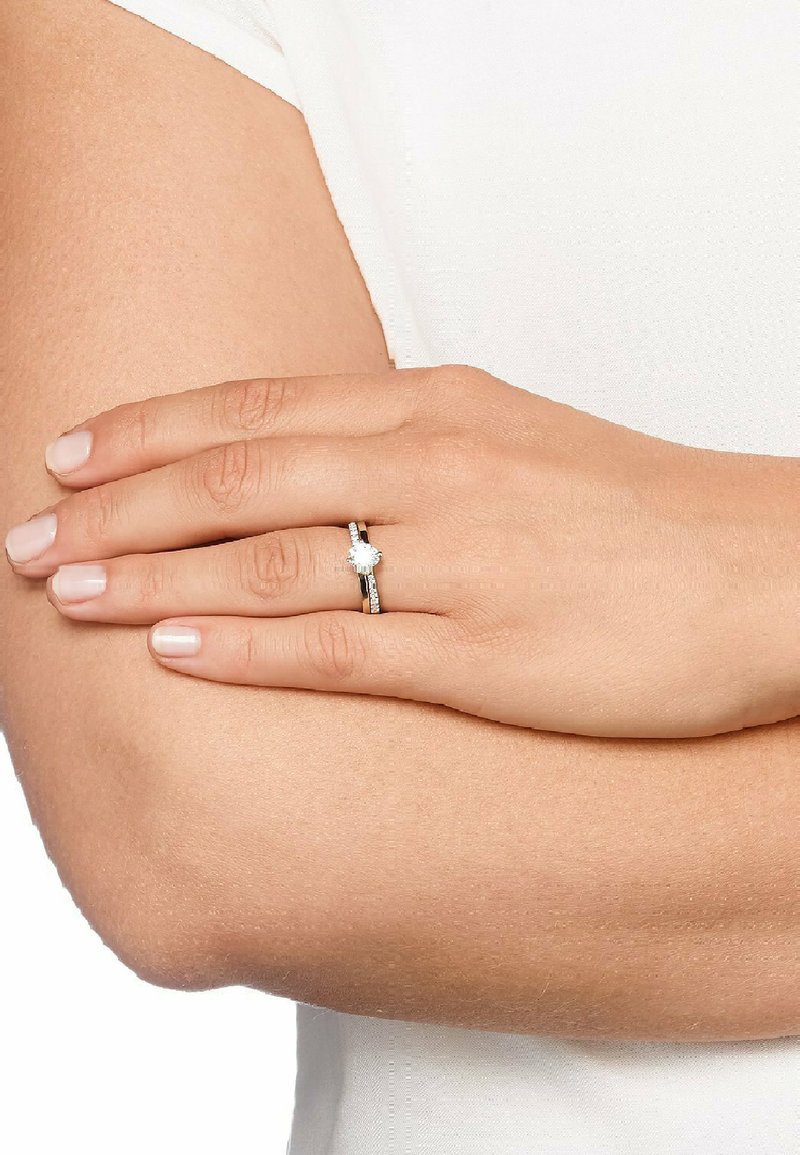 Ring i vitt guld med en rund central diamant omgiven av mindre diamanter, fäst på en polerad ringbåge. Hand som vilar på en arm.