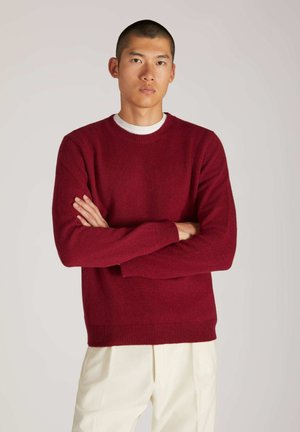 Bordeauxfarbener Strickpullover mit Rundhalsausschnitt, langen Ärmeln sowie gerippten Bündchen und Saum. Kombiniert mit hellen, locker sitzenden Hosen.
