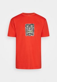 Camiseta de algodón roja con un estampado gráfico de una cara negra con rasgos exagerados, ojos blancos y dientes verdes en el frente.