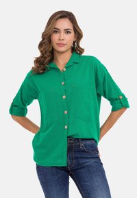 Groene button-up shirt gemaakt van lichte stof, met twee borstzakken, opgerolde mouwen en houten knopen. Gestyled met blauwe jeans.