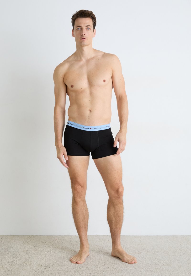 Felsőtest nélküli férfi, aki mezítláb áll szőnyeggel borított padlón, fekete boxeralsót visel világoskék Tommy Hilfiger derékpánttal, fehér fal előtt.