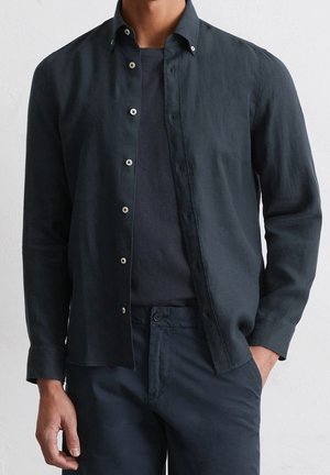 Man draagt een donker losgeknoopt overhemd met lange mouwen over een donker T-shirt met bijpassende donkere broek, één hand in de zak.