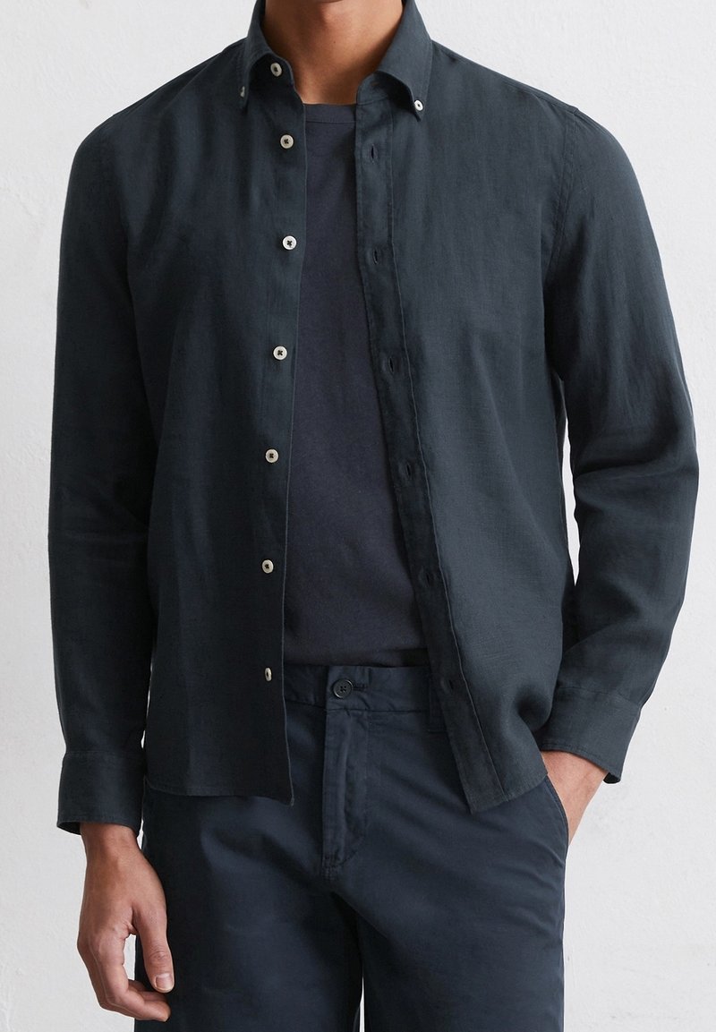 Homme portant une chemise sombre à manches longues déboutonnée par-dessus un t-shirt sombre avec un pantalon sombre assorti, une main dans la poche.