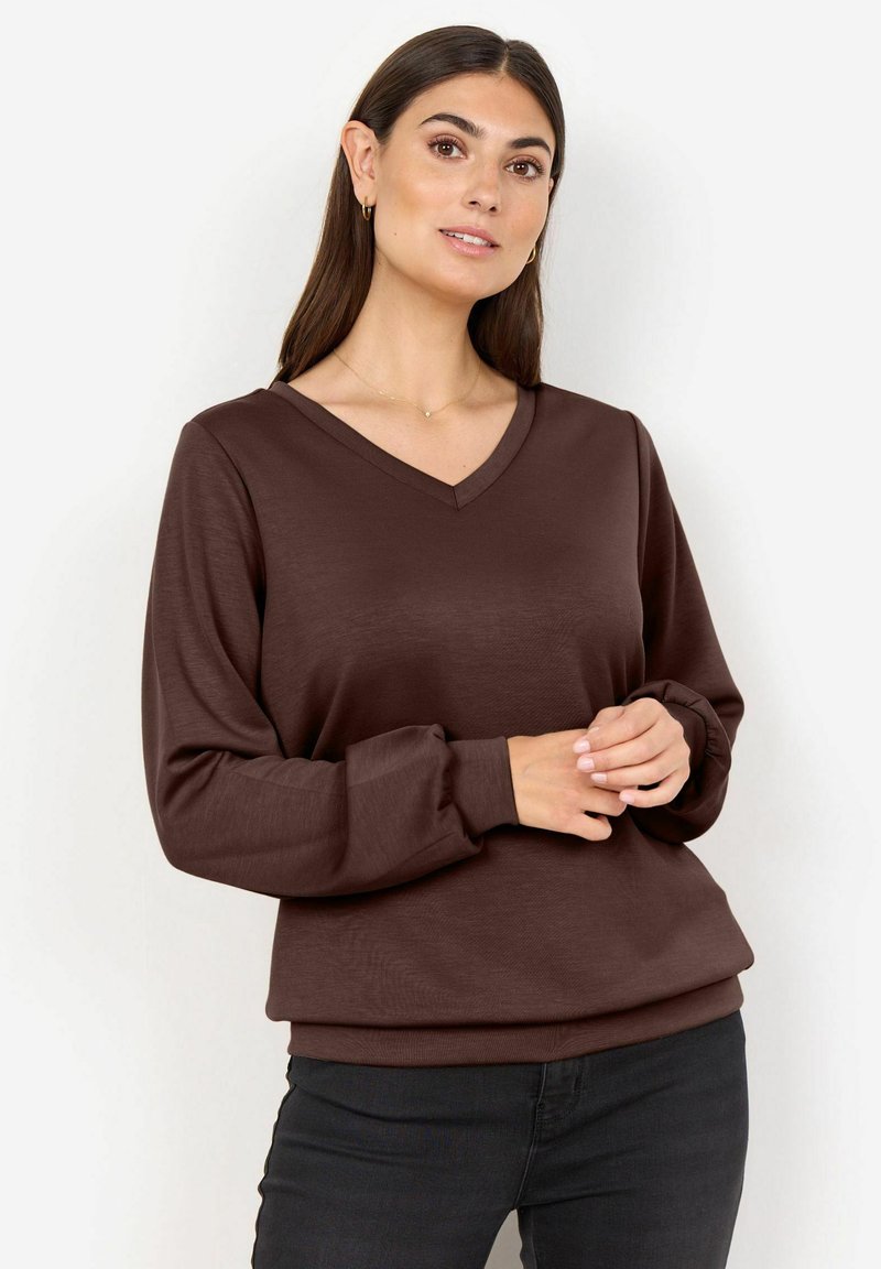 Mujer con cabello largo y oscuro que lleva una blusa marrón de manga larga con escote en V y pantalones negros, de pie con las manos entrelazadas al frente.