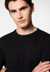 Czarny sweter z prążkowanego dzianiny z okrągłym dekoltem, z długimi rękawami i teksturowaną powierzchnią, noszony przez osobę z rękami skrzyżowanymi.