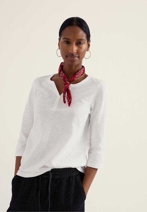 Žena s tmavými vlasmi zviazanými dozadu, oblečená v bielej dlhorukávovej košeli, červenom šatke na krk, zlatých kruhových náušniciach a čiernych nohaviciach s rukami v vreckách.