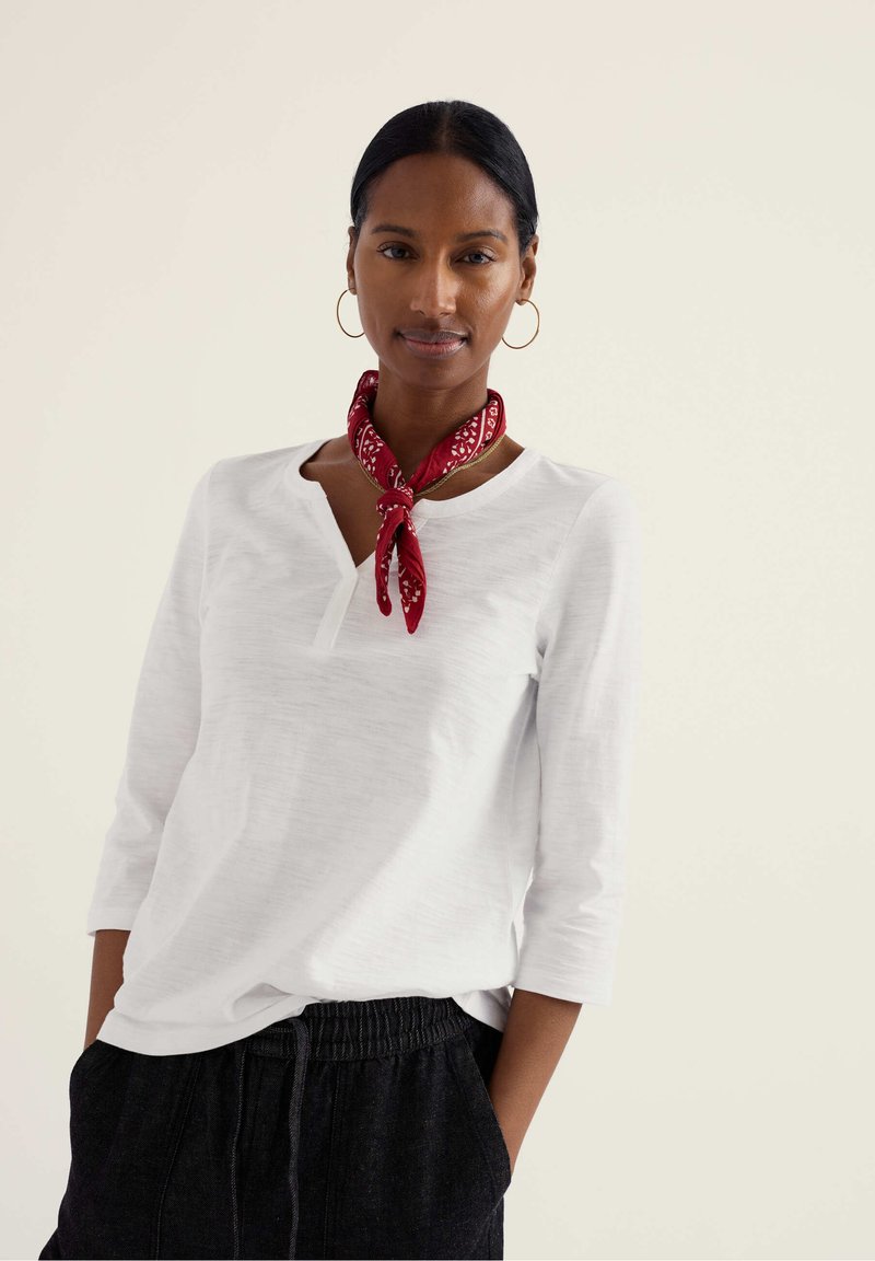 Sieviete ar tumšiem matiem, sasietiem aizmugurē, tērpusies baltā garroku krekliņā, ar sarkanām lakatiņa šalles drapējumu, zelta lokveida auskariem un melnām biksēm ar rokām kabatās.