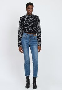Donna in piedi che indossa un maglione a maniche lunghe a righe nere e grigie, jeans blu a vita alta con orli sfrangiati e stivaletti neri.