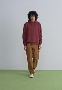 Maroon hoodie with a front pocket, paired with light brown pants. White sneakers complete the outfit. Plain background and green floor.