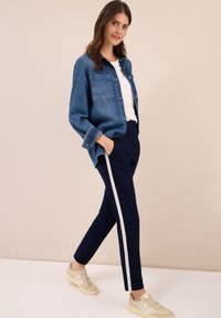 Donna che indossa una giacca di jeans blu, una camicia bianca, pantaloni navy con righe laterali bianche e sneakers beige, in piedi con le mani nelle tasche.