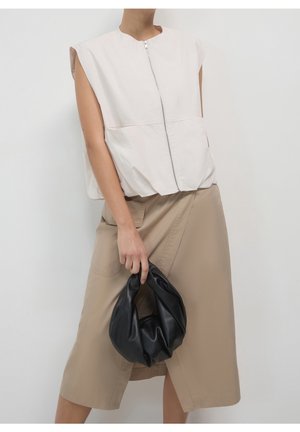 Ærmeløs hvid top med lynlås foran kombineret med en beige omslagsskjørt, holder en lille sort lædertaske med folder i hoften.