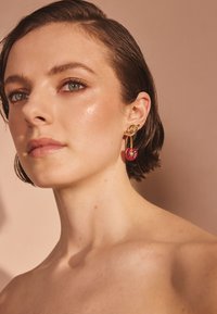 Náušnice v zlatej tónine so srdcovým červeným drahokamom a listovým akcentom. Hladký povrch, moderný dizajn na neutrálom pozadí.