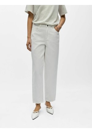 Femme portant un pantalon blanc droit taille haute, une chemise à manches courtes gris clair, des ballerines blanches à bouts pointus, debout avec la main droite dans la poche.