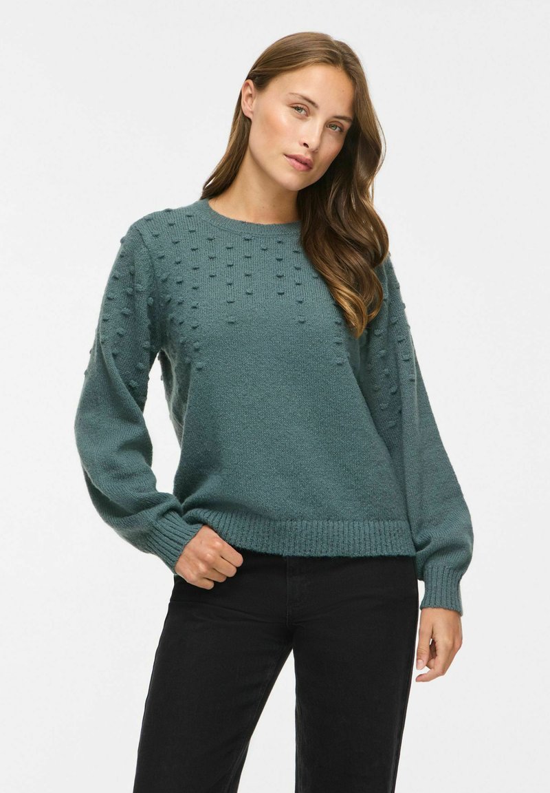 Femme aux longs cheveux bruns portant un pull en maille texturée bleu sarcelle et un pantalon noir, debout sur fond blanc.